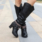 Black knee high boots featuring studded straps and a block heel, crafted from slouchy vegan leather for a stylish and comfortable fit for women. 8