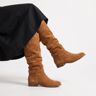 Knee high black boots for women featuring a slouch silhouette and buckled ankle tab, crafted from vegan materials with a low stacked heel. 4